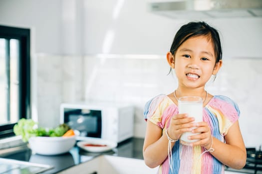 Adorable Asian child holds milk cup in kitchen. Cute girl smiles joyfully savoring the drink. Portrait of happy daughter at home enjoying calcium-rich liquid radiating happiness give me.