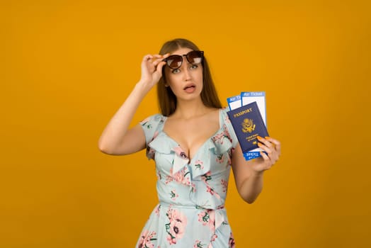 Pop-eyed young woman in blue dress with flowers and sunglasses is holding airline tickets with a passport on a yellow background. Rejoices in the resumption of tourism after the coronovirus pandemic.