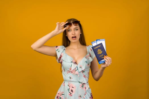 Pop-eyed young woman in blue dress with flowers and sunglasses is holding airline tickets with a passport on a yellow background. Rejoices in the resumption of tourism after the coronovirus pandemic.