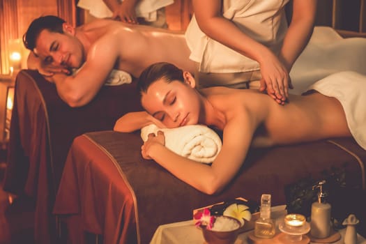 Caucasian couple customer enjoying relaxing anti-stress spa massage and pampering with beauty skin recreation leisure in warm candle lighting ambient salon spa at luxury resort or hotel. Quiescent