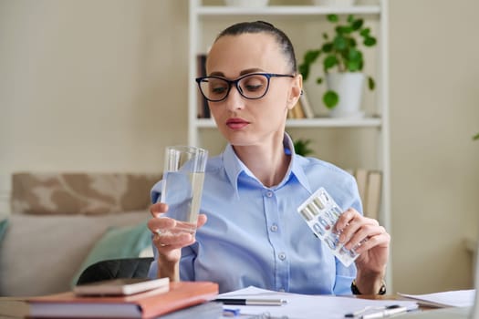 Serious woman at workplace holding blister with capsules glass of water. Female taking painkillers vitamins nutritional supplements antiviral drugs antioxidants antidepressants. Pharmacy health care