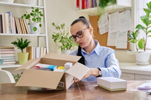 Satisfied female consumer buyer sitting at home unpacking cardboard box with online purchases. Woman unpacking boxes with cosmetics care products vitamins perfumes. Delivery by mail, online store