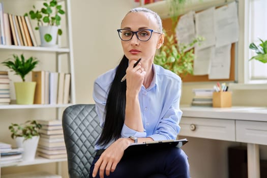 Portrait of confident female psychotherapist with clipboard at workplace in office. Professional mental therapist counselor psychologist social worker looking at camera. Health care service, treatment