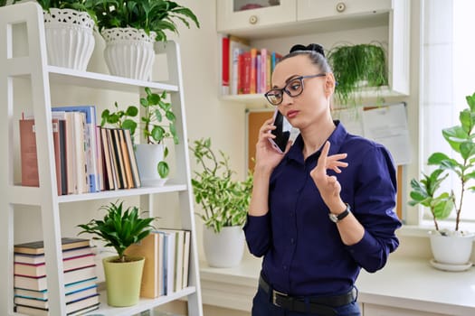 Beautiful emotional woman in her 30s talking on a cell phone, standing in her room at home. Conversation, communication, work, leisure, lifestyle, people concept