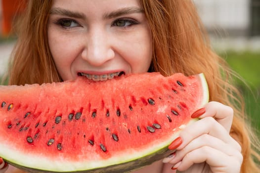 Close-up portrait of red-haired young woman with braces eating watermelon outdoors.
