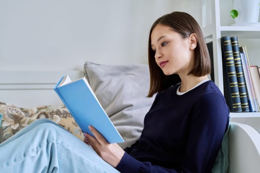 Young woman reading paper book, relaxed sitting on sofa at home. Fiction, hobby, leisure, youth concept