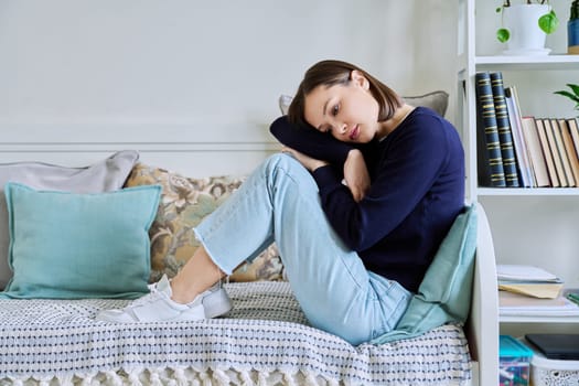 Young sad upset unhappy woman sitting on couch at home. Frustrated confused female experiencing difficulties. Mental problems, feelings, loneliness, stress, depression, difficulties, youth concept