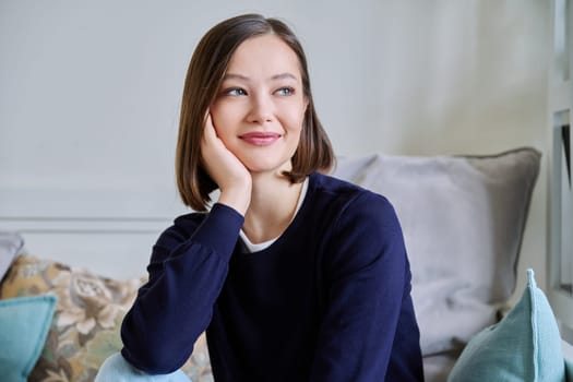 Portrait of young attractive smiling woman in home interior. Happy cheerful relaxed 20s female looking away, sitting on couch. Beauty, youth, happiness, health, lifestyle concept