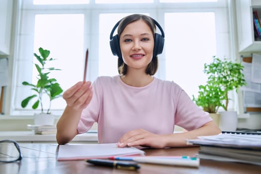 Webcam portrait of young 20s female student wearing headphones, sitting at home at desk, looking at camera, talking. Internet online webinar, video chat call conference, technology education training