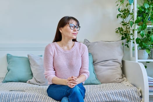 Portrait of calm, thoughtful, relaxed 40s woman, female looking out window sitting on couch, bad at home. Middle age, mental health, lifestyle, people concept