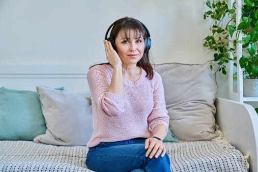 Middle-aged woman in headphones listening to audio, music, audiobook, communication, relaxed vacationer sitting on couch at home