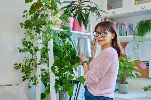 Middle-aged woman watering pots with plants from watering can, home interior. Smiling female looking at camera. Home landscaping, eco green trends, lifestyle concept