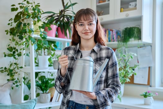 Young teenage woman watering pots with plants from watering can, home interior. Smiling girl looking at camera. Home landscaping, eco green trends, lifestyle concept