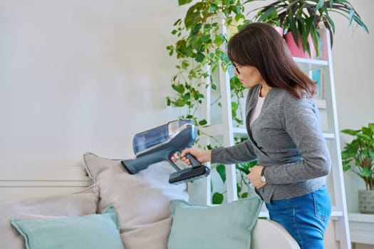 Middle aged woman doing vacuuming, vacuuming pillows on couch. Cleanliness, routine house cleaning, home hygiene, housecleaning, housekeeping, housework concept