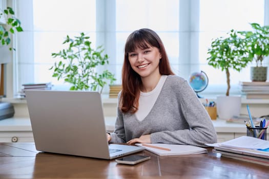 Young female college student studying at home at desk using computer laptop, writing in notebook, smiling looking at camera. E-learning, education, technology, knowledge, youth concept