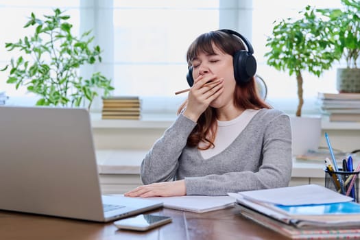 Young female student teenager in headphones yawning covering mouth with hand, bored sleepy tired female sitting at table with computer laptop