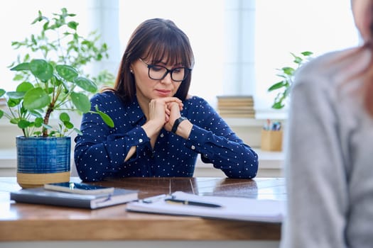 Mature sad woman in therapy session with mental professional psychologist, sitting at table in office of therapist counselor social worker. Psychology psychotherapy mental assistance treatment support