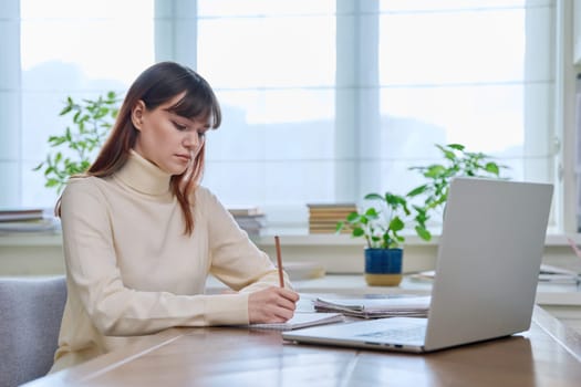 College student girl sitting at desk using laptop computer, making notes in study notebook, at home. Teenager female watching webinar, preparing for exam tests, studying remotely.