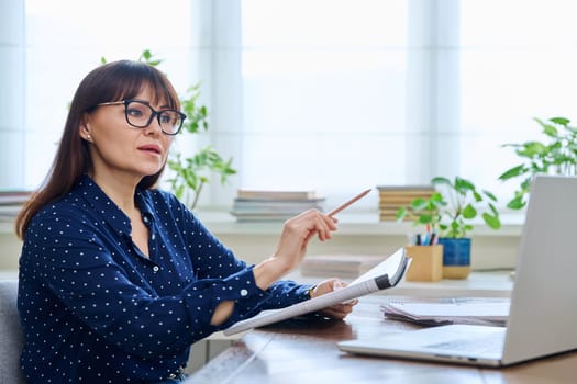 Middle aged woman teacher with textbook working online using laptop computer in home office. Video conference remote meeting consultation e lesson training education technology concept