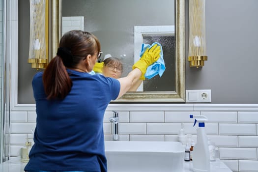 Woman cleaning bathroom, washing mirror with spray bottle and professional washcloth for cleaning glass without streaks. Housekeeping housework housecleaning cleaning service concept
