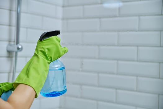 Close-up hands in gloves cleaning bathroom, washing glass in shower, with detergent spray and professional rag, white tiles copy space. Housekeeping, housework, housecleaning, cleaning service concept