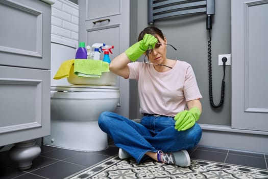 Tired woman sitting on the floor after cleaning bathroom, toilet. Routine house cleaning, home hygiene, housecleaning service, housekeeping, housework