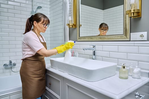 Woman in apron with detergent washcloth cleaning in bathroom, washing sink washbasin. Housewife cleaning house, cleaner service worker at workplace. Home hygiene housecleaning housekeeping housework