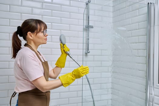 Middle-aged woman in apron, gloves cleaning bathroom, cleans glass in shower. Female housewife cleaning house, service worker at workplace. Home hygiene, housecleaning service, housekeeping, housework