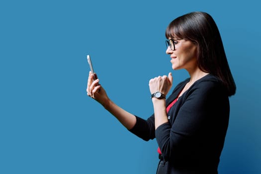 Profile view middle aged smiling woman using smartphone on blue background. Mature female looking at phone in hands. Technologies mobile apps applications internet work business leisure communication