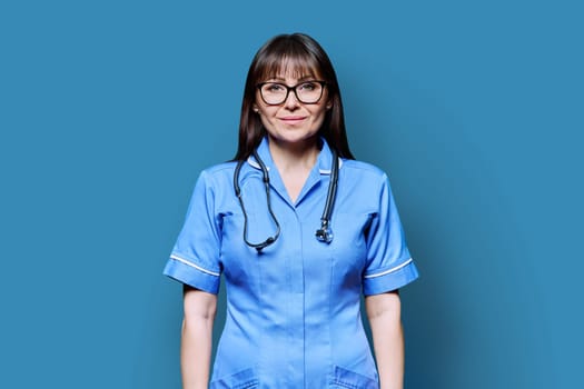 Portrait of smiling middle-aged woman nurse in blue with stethoscope on blue studio background. Medical services, nhs, health, professional assistance, medical care concept