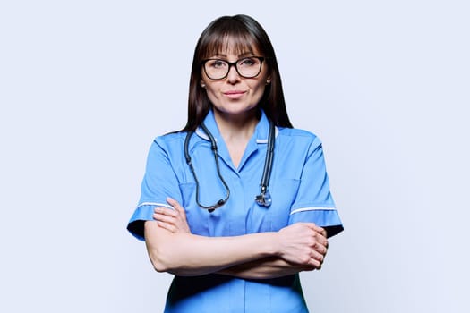 Portrait of serious middle-aged woman nurse with crossed arms in blue with stethoscope on white studio background. Medical services, nhs, health, professional assistance, medical care concept