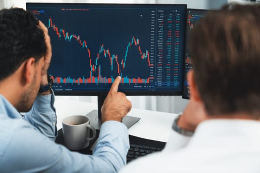 Investor stock trading with serious face sharing with coworker while drinking coffee by using smartphone and pc to show dynamic database of trend graph. Monitor showing investment exchange. Sellable.