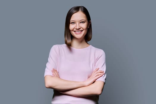 Young beautiful confident woman with crossed arms on gray studio background. Smiling attractive 20s female looking at camera. Lifestyle, fashion beauty style, youth business work education concept