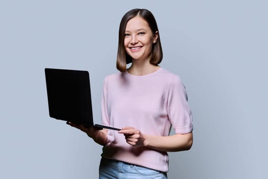 Smiling young woman with laptop computer on grey studio background. Successful attractive female looking at camera. Business, work, job, study, education, e-learning, communication, technology concept