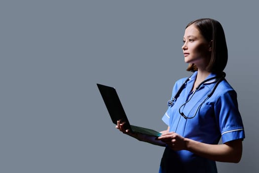 Serious young female nurse using laptop computer, profile view on gray studio background. Mobile apps applications technologies in medical services health professional assistance medical care concept