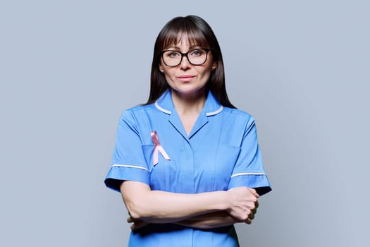 Portrait of serious female nurse in blue uniform with pink ribbon symbolizing International Breast Cancer Day on grey background. Treatment diagnosis of breast cancer, medicine women's health concept