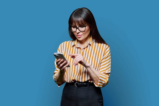 Serious business mature woman using smartphone on blue background. Middle-aged confident successful female holding phone texting reading looking. Mobile internet online technologies apps applications