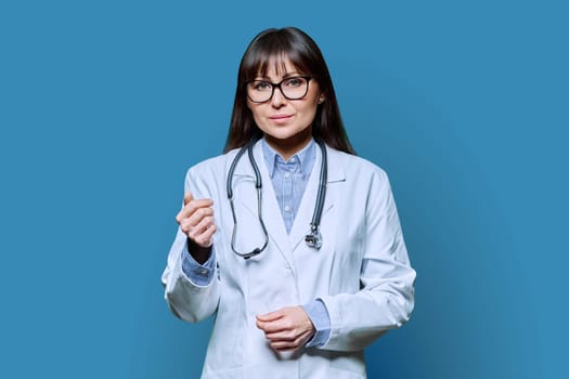 Confident friendly smiling middle-aged female doctor in white lab coat with stethoscope, looking at camera on blue studio background. Healthcare, medicine, staff, treatment, medical services concept