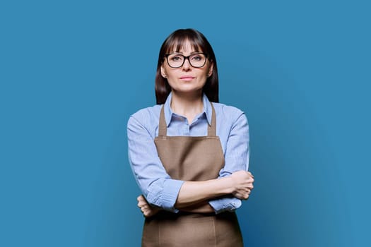 Confident serious middle-aged woman in apron on blue background. Successful mature female small business owner, service worker, entrepreneur looking at camera with crossed arms. Business work people