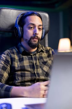 Male individual wearing wireless headphones and looking at his digital laptop. Sitting at a desk is young man utilizing his headset and portable computer for entertainment, watching a movie at home.