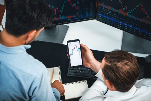 Investor stock officer focusing on dynamic exchange rate database on smartphone, comparing with market dynamic graph with monitor screen. Concept of analysis trading technology investment. Sellable.