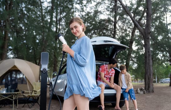 Outdoor adventure and family vacation camping at sea travel by eco friendly car. Cheerful woman or mother holding, pointing EV charger point with playful and happiness posture in campsite. Perpetual