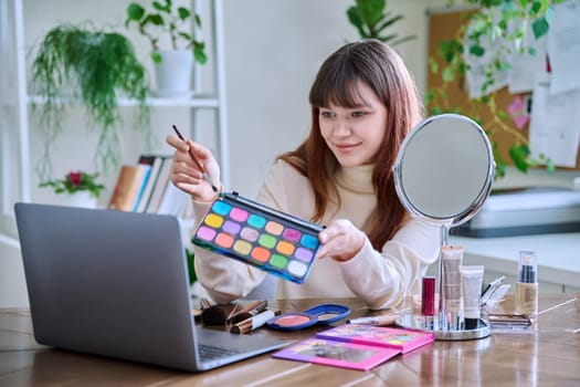 Young female showing cosmetics to laptop webcam, sitting at table at home. Beauty blog vlog, online course, training webinar on professional use of decorative cosmetics