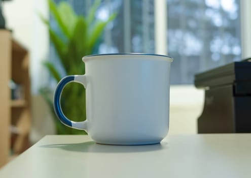 A enamel white mug with a blue handle, a type of drinkware, is placed on a white table, a piece of tableware or serveware