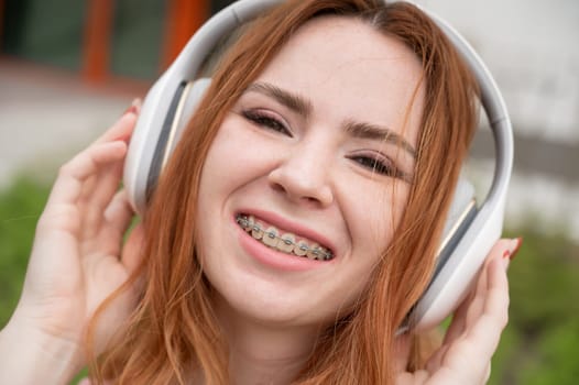 Portrait of a young red-haired woman with braces on her teeth listening to music on headphones outdoors