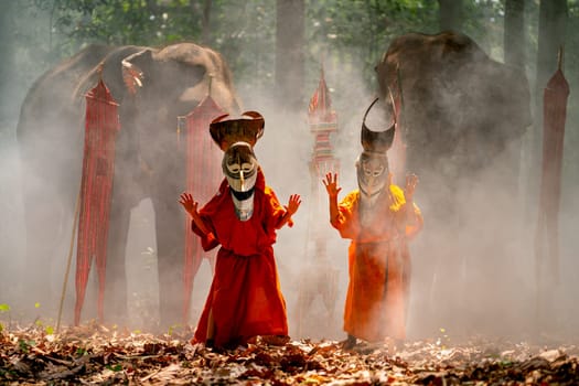 Two boy wear Pee Ta Khon, traditional culture art as ghost of Asian culture, dress and look at camera and elephants stand in the background and they stay in forest.