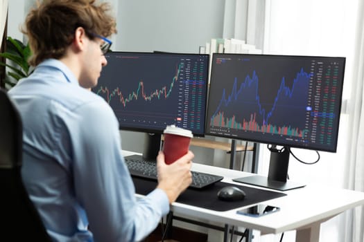 Working young business trader focusing on market stock graph data in two screens, holding coffee cup with back side view at modern office. Concept of analyzing dynamic financial exchange rate. Gusher.