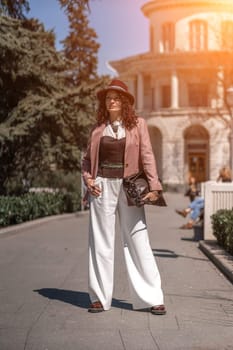 Woman park city. Stylish woman in a hat walks in a park in the city. Dressed in white corset trousers and a pink jacket with a bag in her hands