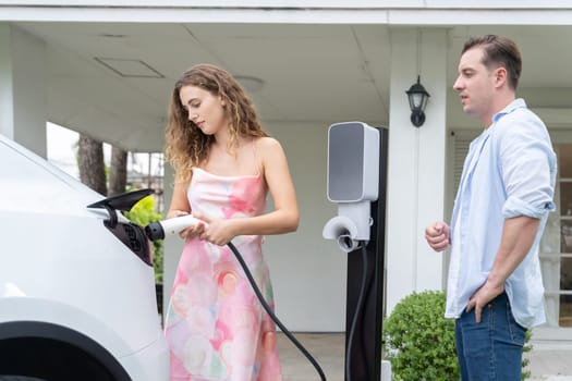 Happy and lovely couple with eco-friendly conscious recharging electric vehicle from EV home charging station. EV car technology utilized for residential home to future sustainability. Synchronos