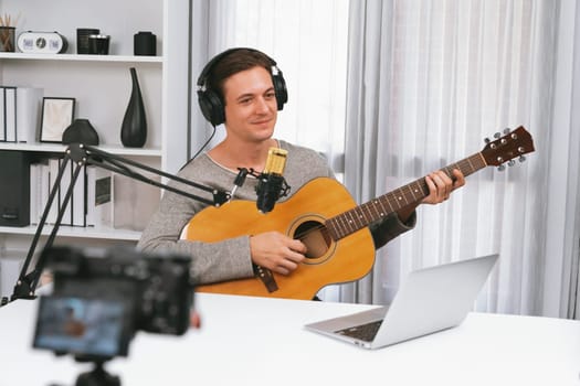 Host channel in smart singer recording by camera, playing guitar along singing, broadcasting on social media channel, wearing headphones to record video streamer at white modern studio. Pecuniary.